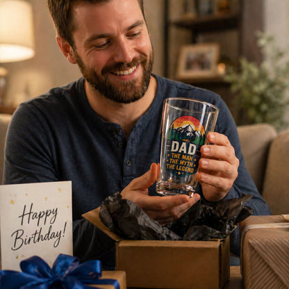 Man holding a personalized glass mug with a 'Dad' message, surrounded by birthday gifts in a home setting.