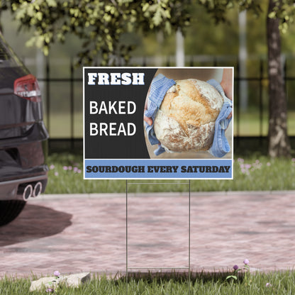 Promotional square sign for fresh baked bread with a loaf of bread and gloved hands, placed outdoors.